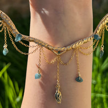 Load image into Gallery viewer, A hand holding up an Intricately hand braided wire wrapped necklace made with Gold Plated Wire lined with blue tourmaline gemstones, Gold Plated Diamond Cut chain festoons with dripping blue tourmaline accent gems, and finished with a gold wire wrapped pear shaped blue topaz pendant with swirly accents, against greenery in the background.