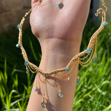 Load image into Gallery viewer, A hand holding up an Intricately hand braided wire wrapped necklace made with Gold Plated Wire lined with blue tourmaline gemstones, Gold Plated Diamond Cut chain festoons with dripping blue tourmaline accent gems, and finished with a gold wire wrapped pear shaped blue topaz pendant with swirly accents, against greenery in the background.