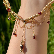 Load image into Gallery viewer, A hand holding up an Intricately hand braided wire wrapped necklace made with Gold Plated Wire lined with red garnet gemstones, Gold Plated Diamond Cut chain festoons with dripping red garnet accent gems, and finished with a gold wire wrapped pear shaped red garnet pendant with swirly accents, against greenery in the background.