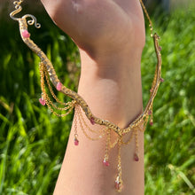 Load image into Gallery viewer, A hand holding up an Intricately hand braided wire wrapped necklace made with Gold Plated Wire lined with pink tourmaline gemstones, Gold Plated Diamond Cut chain festoons with dripping pink tourmaline accent gems, and finished with a gold wire wrapped pear shaped pink tourmaline pendant with swirly accents, against greenery in the background.