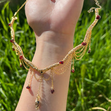 Load image into Gallery viewer, A hand holding up an Intricately hand braided wire wrapped necklace made with Gold Plated Wire lined with red garnet gemstones, Gold Plated Diamond Cut chain festoons with dripping red garnet accent gems, and finished with a gold wire wrapped pear shaped red garnet pendant with swirly accents, against greenery in the background.
