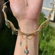 Load image into Gallery viewer, A hand holding up an Intricately hand braided wire wrapped necklace made with Gold Plated Wire lined with blue apatite gemstones, Gold Plated Diamond Cut chain festoons with dripping blue apatite accent gems, and finished with a gold wire wrapped oval shaped blue aquamarine pendant with swirly accents, against greenery in the background.