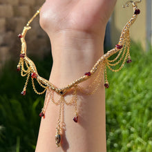 Load image into Gallery viewer, A hand holding up an Intricately hand braided wire wrapped necklace made with Gold Plated Wire lined with red garnet gemstones, Gold Plated Diamond Cut chain festoons with dripping red garnet accent gems, and finished with a gold wire wrapped pear shaped red garnet pendant with swirly accents, against greenery in the background.