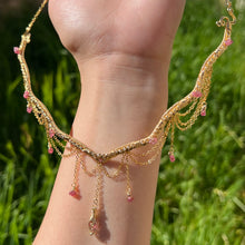 Load image into Gallery viewer, a close up of a woman in a pink dress showcasing an Intricately hand braided wire wrapped necklace made with Gold Plated Wire lined with pink tourmaline gemstones, Gold Plated Diamond Cut chain festoons with dripping pink tourmaline accent gems, and finished with a gold wire wrapped pear shaped pink tourmaline pendant with swirly accents, against greenery in the background.