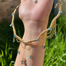 Load image into Gallery viewer, A hand holding up an Intricately hand braided wire wrapped necklace made with Gold Plated Wire lined with blue tourmaline gemstones, Gold Plated Diamond Cut chain festoons with dripping blue tourmaline accent gems, and finished with a gold wire wrapped pear shaped blue topaz pendant with swirly accents, against greenery in the background.