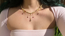 Load image into Gallery viewer, a close up of a woman in a pink dress showcasing an Intricately hand braided wire wrapped necklace made with Gold Plated Wire lined with red garnet gemstones, Gold Plated Diamond Cut chain festoons with dripping red garnet accent gems, and finished with a gold wire wrapped pear shaped red garnet pendant with swirly accents, against greenery in the background.