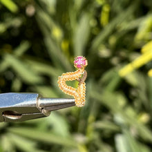Load image into Gallery viewer, intricately hand wire wrapped gold Vipera Serpent Pierceless Nose Cuff with a prong set round cut ruby displayed on a metal holder against a blurred greenery background.