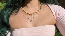 Load image into Gallery viewer, a close up of a woman in a pink dress showcasing an Intricately hand braided wire wrapped necklace made with Gold Plated Wire lined with blue tourmaline gemstones, Gold Plated Diamond Cut chain festoons with dripping blue tourmaline accent gems, and finished with a gold wire wrapped pear shaped blue topaz pendant with swirly accents, against greenery in the background.
