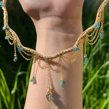 Load image into Gallery viewer, A hand holding up an Intricately hand braided wire wrapped necklace made with Gold Plated Wire lined with blue tourmaline gemstones, Gold Plated Diamond Cut chain festoons with dripping blue tourmaline accent gems, and finished with a gold wire wrapped pear shaped blue topaz pendant with swirly accents, against greenery in the background.