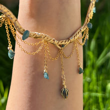 Load image into Gallery viewer, A hand holding up an Intricately hand braided wire wrapped necklace made with Gold Plated Wire lined with blue tourmaline gemstones, Gold Plated Diamond Cut chain festoons with dripping blue tourmaline accent gems, and finished with a gold wire wrapped pear shaped blue topaz pendant with swirly accents, against greenery in the background.