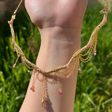 Load image into Gallery viewer, A hand holding up an Intricately hand braided wire wrapped necklace made with Gold Plated Wire lined with pink tourmaline gemstones, Gold Plated Diamond Cut chain festoons with dripping pink tourmaline accent gems, and finished with a gold wire wrapped pear shaped pink tourmaline pendant with swirly accents, against greenery in the background.