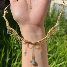Load image into Gallery viewer, A hand holding up an Intricately hand braided wire wrapped necklace made with Gold Plated Wire lined with blue apatite gemstones, Gold Plated Diamond Cut chain festoons with dripping blue apatite accent gems, and finished with a gold wire wrapped oval shaped blue aquamarine pendant with swirly accents, against greenery in the background.