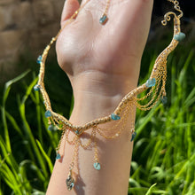 Load image into Gallery viewer, A hand holding up an Intricately hand braided wire wrapped necklace made with Gold Plated Wire lined with blue tourmaline gemstones, Gold Plated Diamond Cut chain festoons with dripping blue tourmaline accent gems, and finished with a gold wire wrapped pear shaped blue topaz pendant with swirly accents, against greenery in the background.