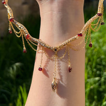 Load image into Gallery viewer, A hand holding up an Intricately hand braided wire wrapped necklace made with Gold Plated Wire lined with red garnet gemstones, Gold Plated Diamond Cut chain festoons with dripping red garnet accent gems, and finished with a gold wire wrapped pear shaped red garnet pendant with swirly accents, against greenery in the background.