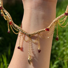 Load image into Gallery viewer, A hand holding up an Intricately hand braided wire wrapped necklace made with Gold Plated Wire lined with red garnet gemstones, Gold Plated Diamond Cut chain festoons with dripping red garnet accent gems, and finished with a gold wire wrapped pear shaped red garnet pendant with swirly accents, against greenery in the background.