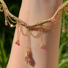 Load image into Gallery viewer, A hand holding up an Intricately hand braided wire wrapped necklace made with Gold Plated Wire lined with pink tourmaline gemstones, Gold Plated Diamond Cut chain festoons with dripping pink tourmaline accent gems, and finished with a gold wire wrapped pear shaped pink tourmaline pendant with swirly accents, against greenery in the background.