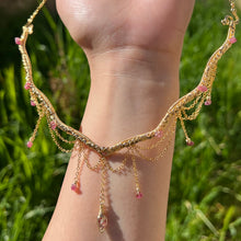 Load image into Gallery viewer, A hand holding up an Intricately hand braided wire wrapped necklace made with Gold Plated Wire lined with pink tourmaline gemstones, Gold Plated Diamond Cut chain festoons with dripping pink tourmaline accent gems, and finished with a gold wire wrapped pear shaped pink tourmaline pendant with swirly accents, against greenery in the background.