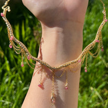 Load image into Gallery viewer, a close up of a woman in a pink dress showcasing an Intricately hand braided wire wrapped necklace made with Gold Plated Wire lined with pink tourmaline gemstones, Gold Plated Diamond Cut chain festoons with dripping pink tourmaline accent gems, and finished with a gold wire wrapped pear shaped pink tourmaline pendant with swirly accents, against greenery in the background.