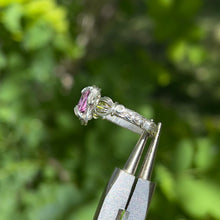 Load image into Gallery viewer, A silver wire wrapped ring featuring a pear cut pink tourmaline center gemstone and round peridot cabochon side stones, with intricate hand braided wire wrapped band details, displayed against a plant background.