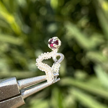 Load image into Gallery viewer, intricately hand wire wrapped silver Vipera Serpent Pierceless Nose Cuff with a prong set red garnet cabochon displayed on a metal holder against a blurred greenery background.