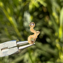 Load image into Gallery viewer, intricately hand wire wrapped gold Vipera Serpent Pierceless Nose Cuff with a prong set rose de France purple amethyst cabochon displayed on a metal holder against a blurred greenery background.