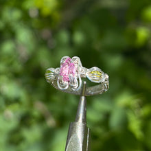 Load image into Gallery viewer, A silver wire wrapped ring featuring a pear cut pink tourmaline center gemstone and round peridot cabochon side stones, with intricate hand braided wire wrapped band details, displayed against a plant background.