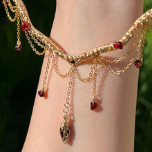 Load image into Gallery viewer, A hand holding up an Intricately hand braided wire wrapped necklace made with Gold Plated Wire lined with red garnet gemstones, Gold Plated Diamond Cut chain festoons with dripping red garnet accent gems, and finished with a gold wire wrapped pear shaped red garnet pendant with swirly accents, against greenery in the background.