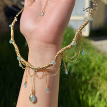 Load image into Gallery viewer, A hand holding up an Intricately hand braided wire wrapped necklace made with Gold Plated Wire lined with blue apatite gemstones, Gold Plated Diamond Cut chain festoons with dripping blue apatite accent gems, and finished with a gold wire wrapped oval shaped blue aquamarine pendant with swirly accents, against greenery in the background.