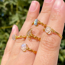 Load image into Gallery viewer, A selection of gold-toned rings featuring moonstone or labradorite stones, with wire crescent moons around them displayed on a person's fingers.