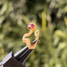 Load image into Gallery viewer, intricately hand wire wrapped gold Vipera Serpent Pierceless Nose Cuff with a prong set red garnet cabochon displayed on a metal holder against a blurred greenery background.