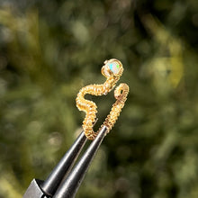Load image into Gallery viewer, intricately hand wire wrapped gold Vipera Serpent Pierceless Nose Cuff with a prong set round white ethiopian welo opal cabochon displayed on a metal holder against a blurred greenery background.