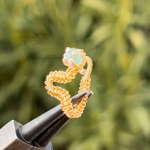 Load image into Gallery viewer, intricately hand wire wrapped gold Vipera Serpent Pierceless Nose Cuff with a prong set raw cut blue apatite chip displayed on a metal holder against a blurred greenery background.