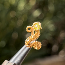 Load image into Gallery viewer, intricately hand wire wrapped gold Vipera Serpent Pierceless Nose Cuff with a prong set green peridot cabochon displayed on a metal holder against a blurred greenery background.