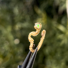 Load image into Gallery viewer, intricately hand wire wrapped gold Vipera Serpent Pierceless Nose Cuff with a prong set round white ethiopian welo opal cabochon displayed on a metal holder against a blurred greenery background.