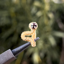 Load image into Gallery viewer, intricately hand wire wrapped gold Vipera Serpent Pierceless Nose Cuff with a prong set black Ethiopian welo opal cabochon displayed on a metal holder against a blurred greenery background.