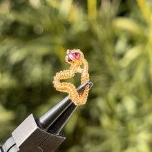 Load image into Gallery viewer, intricately hand wire wrapped gold Vipera Serpent Pierceless Nose Cuff with a prong set red garnet cabochon displayed on a metal holder against a blurred greenery background.
