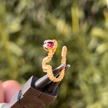 Load image into Gallery viewer, intricately hand wire wrapped gold Vipera Serpent Pierceless Nose Cuff with a prong set red garnet cabochon displayed on a metal holder against a blurred greenery background.