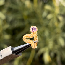 Load image into Gallery viewer, intricately hand wire wrapped gold Vipera Serpent Pierceless Nose Cuff with a prong set purple amethyst cabochon displayed on a metal holder against a blurred greenery background.