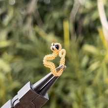 Load image into Gallery viewer, intricately hand wire wrapped gold Vipera Serpent Pierceless Nose Cuff with a prong set black onyx cabochon displayed on a metal holder against a blurred greenery background.
