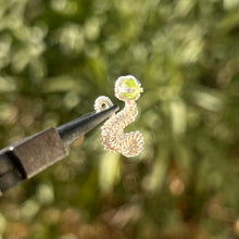 Load image into Gallery viewer, intricately hand wire wrapped silver Vipera Serpent Pierceless Nose Cuff with a prong set green peridot cabochon displayed on a metal holder against a blurred greenery background.