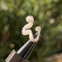 Load image into Gallery viewer, intricately hand wire wrapped silver Vipera Serpent Pierceless Nose Cuff with a prong set white rainbow moonstone cabochon displayed on a metal holder against a blurred greenery background.