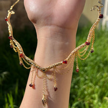 Load image into Gallery viewer, A hand holding up an Intricately hand braided wire wrapped necklace made with Gold Plated Wire lined with red garnet gemstones, Gold Plated Diamond Cut chain festoons with dripping red garnet accent gems, and finished with a gold wire wrapped pear shaped red garnet pendant with swirly accents, against greenery in the background.
