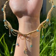 Load image into Gallery viewer, A hand holding up an Intricately hand braided wire wrapped necklace made with Gold Plated Wire lined with blue tourmaline gemstones, Gold Plated Diamond Cut chain festoons with dripping blue tourmaline accent gems, and finished with a gold wire wrapped pear shaped blue topaz pendant with swirly accents, against greenery in the background.
