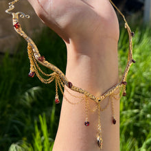 Load image into Gallery viewer, A hand holding up an Intricately hand braided wire wrapped necklace made with Gold Plated Wire lined with red garnet gemstones, Gold Plated Diamond Cut chain festoons with dripping red garnet accent gems, and finished with a gold wire wrapped pear shaped red garnet pendant with swirly accents, against greenery in the background.
