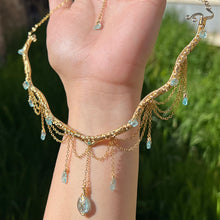 Load image into Gallery viewer, A hand holding up an Intricately hand braided wire wrapped necklace made with Gold Plated Wire lined with blue apatite gemstones, Gold Plated Diamond Cut chain festoons with dripping blue apatite accent gems, and finished with a gold wire wrapped oval shaped blue aquamarine pendant with swirly accents, against greenery in the background.
