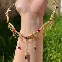 Load image into Gallery viewer, A hand holding up an Intricately hand braided wire wrapped necklace made with Gold Plated Wire lined with red garnet gemstones, Gold Plated Diamond Cut chain festoons with dripping red garnet accent gems, and finished with a gold wire wrapped pear shaped red garnet pendant with swirly accents, against greenery in the background.
