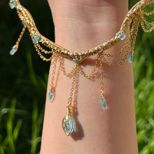 Load image into Gallery viewer, A hand holding up an Intricately hand braided wire wrapped necklace made with Gold Plated Wire lined with blue apatite gemstones, Gold Plated Diamond Cut chain festoons with dripping blue apatite accent gems, and finished with a gold wire wrapped oval shaped blue aquamarine pendant with swirly accents, against greenery in the background.
