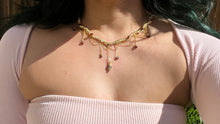 Load image into Gallery viewer, a close up of a woman in a pink dress showcasing an Intricately hand braided wire wrapped necklace made with Gold Plated Wire lined with red garnet gemstones, Gold Plated Diamond Cut chain festoons with dripping red garnet accent gems, and finished with a gold wire wrapped pear shaped red garnet pendant with swirly accents, against greenery in the background.
