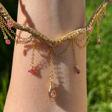 Load image into Gallery viewer, A hand holding up an Intricately hand braided wire wrapped necklace made with Gold Plated Wire lined with pink tourmaline gemstones, Gold Plated Diamond Cut chain festoons with dripping pink tourmaline accent gems, and finished with a gold wire wrapped pear shaped pink tourmaline pendant with swirly accents, against greenery in the background.
