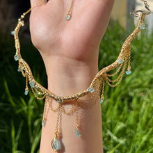 Load image into Gallery viewer, A hand holding up an Intricately hand braided wire wrapped necklace made with Gold Plated Wire lined with blue apatite gemstones, Gold Plated Diamond Cut chain festoons with dripping blue apatite accent gems, and finished with a gold wire wrapped oval shaped blue aquamarine pendant with swirly accents, against greenery in the background.
